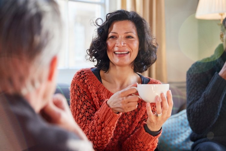 A midlife woman is talking with friends while drinking from a mug.