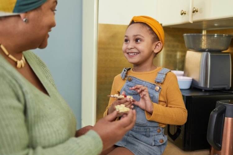 A woman stands while a young girl sits on the kitchen counter. They are smiling and sharing a snack, exemplifying inherent self-worth in midlife.