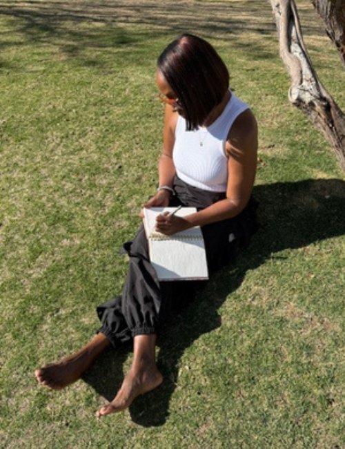 Dr. Arlonda Stevens, author of I Forgot to Put My Mask On First, sits on the grass beside a tree while writing in a journal.